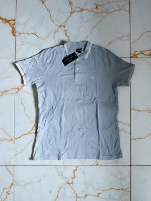 Ljusblå pikétröja från Fred Perry - Snygg ljusblå pikétröja från Fred Perry med klassisk krage och knappar framtill. Tröjan har vita detaljer på krage och ärmslut samt den ikoniska Fred Perry-loggan broderad på bröstet. Perfekt till jeans eller shorts för en clean look.