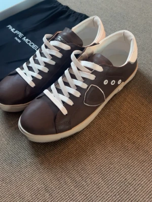 Bruna Philippe Model sneakers - Old money grish.Snygga bruna sneakers från Philippe Model med vita detaljer och vita skosnören. Skorna har en låg silhuett, klassisk gummisula och är tillverkade i läder. Perfekta för dig som gillar stilrena och tidlösa sneakers.