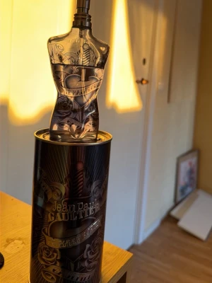 Jean Paul Gaultier Le Male Lover - Unik parfymflaska formad som en manlig torso med tatueringsinspirerat mönster. Flaskan är genomskinlig med mörka detaljer och kommer i en metallburk med matchande design. En cool och ikonisk doft för dig som gillar statement-parfymer. Burken kommer man få med men är lite bränd på toppen för någon har lekt med eld där annars så luktar den väldigt gott med dofter som förklarar sommaren. Denna Doften är Limited edition. Man får också med de ikoniska stället för parfymen 