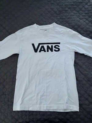 Vit Vans t-shirt med logga - Säljer en klassisk vit t-shirt från Vans med stor svart logga på bröstet. T-shirten har rund hals och korta ärmar, perfekt till en avslappnad streetwear-look. Tillverkad i mjuk bomull för skön känsla hela dagen.