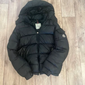 Svart dunjacka från Moncler - Snygg svart dunjacka från Moncler med huva och två dragkedjefickor framtill. Jackan har Moncler-logga på ärmen och quiltad design som ger en puffig look. Perfekt för kalla vinterdagar och riktigt trendig stil.