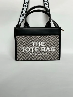 Marc Jacobs The Tote Bag handväska - Säljer en snygg handväska från Marc Jacobs, modell The Tote Bag. Väskan har svart-vitt mönstrad textil på framsidan med tydlig logga, svarta läderdetaljer och både handtag och axelrem med Marc Jacobs-tryck. Perfekt för dig som gillar trendiga accessoarer.