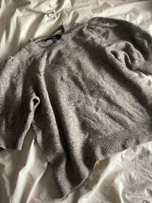 Gråmelerad stickad tröja - Mysig gråmelerad stickad tröja från H&M med rund halsringning och ribbade muddar vid ärmslut och nederkant. Tröjan har en avslappnad passform och puffiga ärmar som ger en trendig look. Perfekt för lager på lager under kyliga dagar.