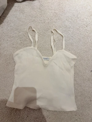 Ribbad vit topp från Zara - Säljer en ribbad vit topp från Zara med smala axelband och en snygg V-ringning. Toppen är croppad och har en figurnära passform, perfekt till jeans eller kjol. Materialet är mjukt och stretchigt för extra komfort. Storlek M men liten i storlek
