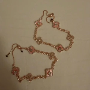 Guldarmband med rosa blommor 1=99 2=150 - Två eleganta armband i guldfärgad metall med blommiga länkar i rosa och vita nyanser. Armbanden har justerbar kedja och detaljer av små pärlor runt blommorna. Perfekt accessoar för att lyfta din stil.🌸🌸