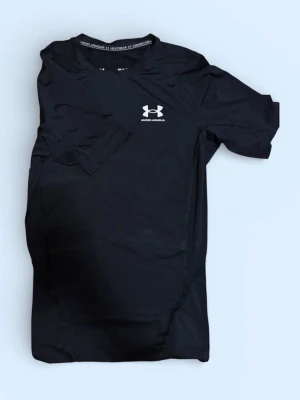 Svart träningströja Under Armour - Svart kortärmad träningströja från Under Armour med logga på bröstet och reflexdetalj i nacken. Tillverkad i ett stretchigt funktionsmaterial som andas, perfekt för intensiva träningspass eller löpning.