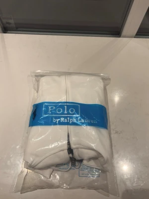 Vit Zip up från Polo Ralph Lauren - Säljer en fräsch vit hoodie från Polo Ralph Lauren med dragkedja framtill och klassisk huva med snören. Hoodien har det ikoniska Polo-logotrycket på bröstet och ribbade muddar vid ärmar och nederkant. Perfekt för en clean och stilren look.