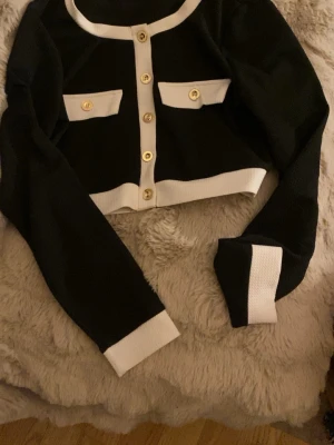 Svart och vit cropped tröja med guldknappar - Trendig svart cropped tröja med vita detaljer och snygga guldknappar framtill. Tröjan har långa ärmar, två dekorativa fickor på bröstet och kontrasterande vita muddar och kanter. Perfekt för en stilren och chic look.