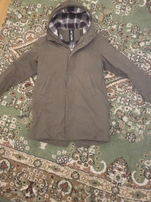 Olivgrön parkas från Replay - Snygg olivgrön parkas från Replay med huva och två snedställda fickor framtill. Insidan har ett rutigt foder i blått, vitt och vinrött samt quiltad nederdel i mörkblått. Perfekt för kyliga dagar och ger en avslappnad streetstil.