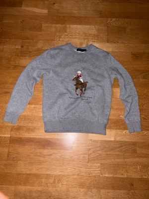 Grå sweatshirt Polo Bear Ralph Lauren - Grå sweatshirt från Ralph Lauren med Polo Bear-motiv på bröstet där björnen rider häst i poloutstyrsel. Rund halsringning och ribbade muddar vid ärmslut och nederkant. Klassisk och avslappnad passform, perfekt till jeans eller mjukiskläder.