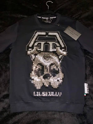 Svart sweatshirt från Philipp Plein - Cool svart sweatshirt från Philipp Plein med ett stort glittrigt döskallemotiv och texten 'LILSKULLY' framtill. Tröjan har rund halsringning, ribbade muddar och en liten logga på ryggen. Tillverkad i mjukt bomullsmaterial för en bekväm känsla.