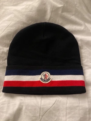 Svart  mössa med ränder - Snygg svart mössa med breda blå, vita och röda ränder längst ner. Framtill sitter klassiska logotyp. Perfekt accessoar för att hålla värmen och addera lite lyx till din outfit.