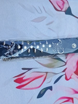 Svart armband med nitar och skorpioner - Säljer ett svart armband i syntet med coola silverfärgade nitar och detaljer i form av skorpioner. Perfekt accessoar för dig som vill sticka ut och addera en edgy vibe till din outfit. Bältet har tryckknappsstängning och flera metallspetsar.