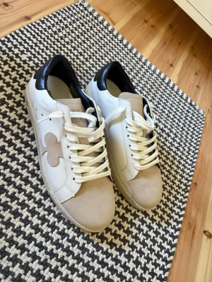 The Gallerist Skor Strl 41, helt oanvända - Snygga sneakers i vitt och beige med svart detalj på hälen och en beige mockadetalj på tån. Skorna har vita snören och en unik beige logga på sidan. Tillverkade i en mix av läder och mocka för en clean och modern look. Låda Ingår.