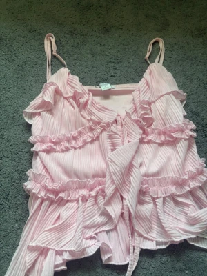 Ljusrosa volang topp med knyt - Supersöt ljusrosa linnetopp med tunna axelband, volanger och knyt framtill. Toppen har ett ribbat tyg och romantiska detaljer som ger en feminin vibe. Perfekt för varma dagar och somriga outfits. 