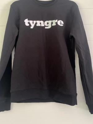Svart sweatshirt med tryck från Tyngre - Svart sweatshirt med vit Tyngre-logga på bröstet. Tröjan har rund halsringning, långa ärmar och en klassisk, avslappnad passform. Perfekt för en sportig och casual stil.