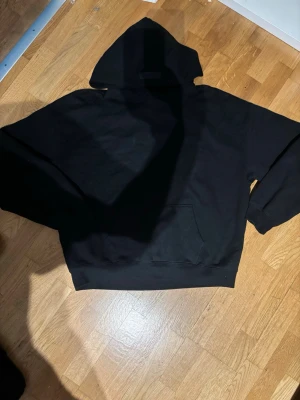 Svart Essentials hoodie med huva - Säljer en svart hoodie från Essentials med stor huva och magficka. Hoodien har en stilren look med diskret Essentials-märke på huvan. Perfekt för dig som gillar streetwear och vill ha en enkel men snygg hoodie.
