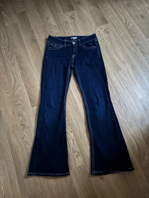 Mörkblå bootcut jeans från Gina  - Storlek 164 Snygga mörkblå bootcut jeans med vita kontrastsömmar och låg midja. Klassisk femficksmodell med detaljerade fickor fram och bak. Jeansen har en lätt utsvängd passform nedtill och är perfekta för en trendig och avslappnad look.