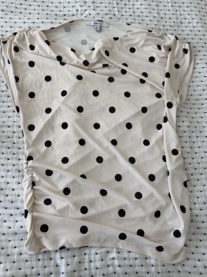 Prickig kortärmad blus från Next - Snygg beige blus med svarta prickar från Next. Blusen har korta ärmar och en lätt draperad halsringning. Materialet är mjukt och stretchigt, perfekt för en avslappnad men trendig look.