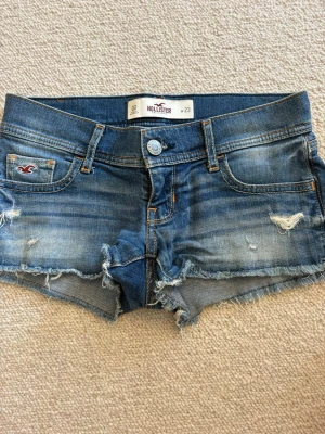 Blå denimshorts från Hollister - Säljer ett par blå denimshorts från Hollister med slitna detaljer och råa kanter. Klassisk femficksmodell med låg midja och knappgylf. Perfekta för varma dagar och ger en avslappnad vibe.💓