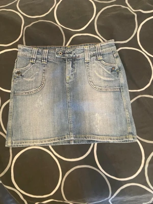 Ljusblå jeanskjol med fickor - Säljer en ljusblå jeanskjol i kort modell med fram- och bakfickor samt bälteshällor. Kjolen har klassisk denimlook och kontrastsömmar. Perfekt till sommaren och enkel att matcha med olika toppar.