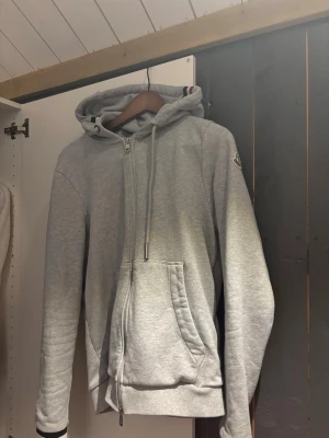 Ljusgrå Moncler hoodie  - Snygg ljusgrå hoodie med dragkedja och huva med dragsko. Fickor framtill och Moncler-logga på ärmen. Huvan har coola ränder i rött, vitt och blått. Perfekt för chill dagar och streetwear vibes.