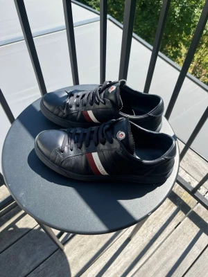 Svarta Moncler skor - Snygga svarta sneakers från Moncler i läder med klassiska röda och vita ränder på sidan samt Moncler-logga på plösen. Skorna har svarta snören och en låg profil som passar till många olika stilar.