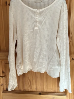 Vit långärmad topp med knappar - Säljer en vit långärmad topp med diskreta ränder och små knappar framtill. Toppen har en lätt ribbad struktur och vågig kant nertill och vid ärmsluten. Perfekt att styla med jeans eller kjol för en enkel och fräsch look.