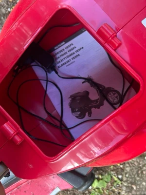 Röd elektrisk leksaksvespa - Säljer en röd elektrisk leksaksvespa med brunt säte och silverfärgade detaljer. Vespan har speglar, handtag och förvaringsutrymme under sätet där laddare och manual ingår. Perfekt för barn som gillar att köra runt och leka.