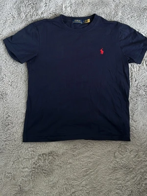 Mörkblå t-shirt från Polo Ralph Lauren - Klassisk mörkblå t-shirt från Polo Ralph Lauren med röd broderad logga på bröstet. T-shirten har rund halsringning och korta ärmar. Tillverkad i mjuk bomull för en skön känsla och enkel stil.