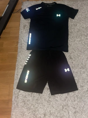 Svart träningsset från Under Armour - Säljer ett svart träningsset från Under Armour med t-shirt och shorts. Båda plaggen har reflekterande loggor och textdetaljer. Perfekt för gymmet eller löpning. Materialet är lätt och andas, vilket gör det bekvämt under träning.