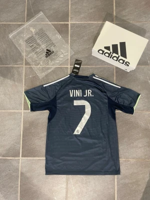 Real Madrid fotbollströja Vini Jr. - Mörkblå Real Madrid fotbollströja från Adidas med VINI JR. och nummer 7 på ryggen. Tröjan har vita detaljer, klubbmärke på bröstet och 'Emirates Fly Better' tryckt framtill. Tillverkad i lätt och ventilerande material, perfekt för fotbollsträning eller match.