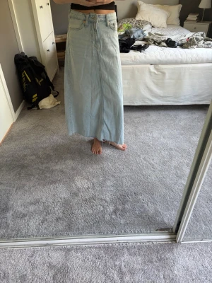 Ljusblå lång jeanskjol - Säljer en ljusblå lång jeanskjol i klassisk denim med femficksmodell och hög midja. Kjolen har en rak passform och slits framtill för extra rörelsefrihet. Perfekt till en trendig vardagslook.