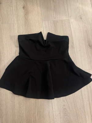 Svart axelbandslös topp - Snygg svart axelbandslös topp med v-formad urringning framtill. Toppen har en utställd kjol och är perfekt för dig som gillar en stilren och trendig look. Tillverkad i ett mjukt och följsamt material.