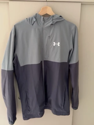 Grå och mörkblå vindjacka Under Armour - Snygg tvåfärgad vindjacka från Under Armour i grått och mörkblått. Jackan har huva, hel dragkedja framtill, bröstficka med dragkedja och två sidofickor. Tillverkad i lätt och vindtätt material, perfekt för blåsiga dagar.