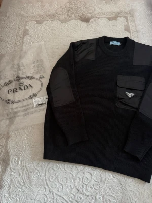 Prada tröja - Säljer en mycket fin och stilren stickad tröja i svart jungfruull med de klassiska Re-Nylon-panelerna på axlarna och armbågarna.Storlek: S (passar även en mindre M) Material: 100% Ull / Detaljer i Re-Nylon Skick: Nyskick, använd vid ett tillfälle. Hör av dig om du vill ha specifika mått eller fler bilder! Skickas spårbart och säkert