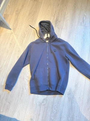Mörkblå CDG zip - Snygg mörkblå hoodie med dragkedja framtill och två stora fickor. Klassisk huva med dragsko och ribbade muddar vid ärmar och nederkant. Liten logga på bröstet. Perfekt för en avslappnad och stilren look.