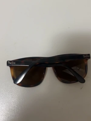 Bruna Wayfarer solglasögon Ray-Ban - Snygga Wayfarer solglasögon från Ray-Ban med brunmelerad båge och mörka glas. Klassisk form som passar till de flesta stilar. Ray-Ban-logga syns på båda sidorna av bågen. Perfekta för soliga dagar.
