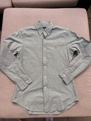 Ralph Lauren skjorta - Klassisk ljusgrön skjorta från Ralph Lauren med button-down krage och broderad logga på bröstet. Skjortan har långa ärmar, vita knappar och är tillverkad i mjuk bomull. Perfekt för en stilren och avslappnad look.