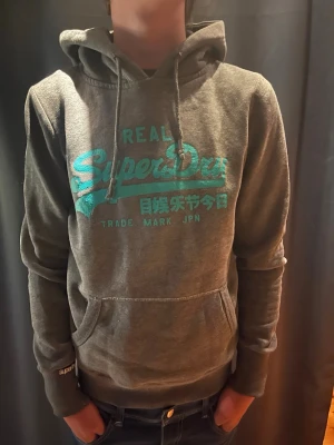 Superdry hoodie - Snygg grå hoodie från Superdry med turkost tryck på bröstet. Huva och stor magficka framtill. Perfekt inför skolan eller bara allmänt. Storlek S, ganska tight 🙌