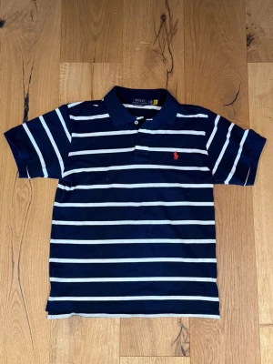 Randig marinblå pikétröja Polo Ralph Lauren - Snygg marinblå pikétröja med vita horisontella ränder från Polo Ralph Lauren. Klassisk krage, korta ärmar och röd broderad logga på bröstet. Perfekt för en stilren och avslappnad look.