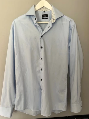 Ljusblå skjorta från Long Island - Snygg ljusblå skjorta från Long Island i modern fit. Skjortan har svarta knappar, lång ärm och en krage med mönstrad insida. Perfekt för en stilren look och enkel att matcha med olika outfits.