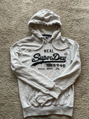 Ljusgrå hoodie från Superdry - Går att pruta. Mycket slitage allt syns inte på bilderna kolla bilderna PM vid frågor. Säljer en ljusgrå hoodie från Superdry med stor logga och text på bröstet. Hoodien har huva med dragsko, känguruficka och ribbade muddar vid ärmar och nederkant. Perfekt för en avslappnad och cool stil.