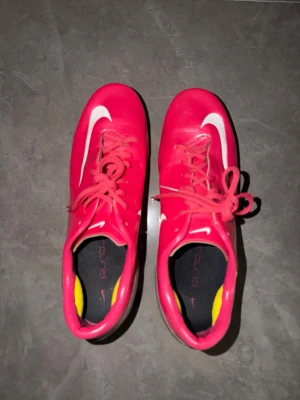Rosa Nike fotbollsskor med dobbar - Säljer ett par snygga rosa fotbollsskor från Nike med vita detaljer och klassisk swoosh på sidan. Skorna har snörning och dobbar för bra grepp på planen. Insidan är svart med gula detaljer. Perfekta för dig som vill sticka ut på fotbollsplanen!