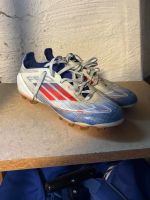 Blåvita fotbollsskor med röda detaljer - Säljer ett par snygga fotbollsskor i blått och vitt med röda detaljer och snörning. Skorna har en sportig design med låg profil och mönstrad sula för bra grepp på planen. Perfekta för dig som vill sticka ut på fotbollen! 