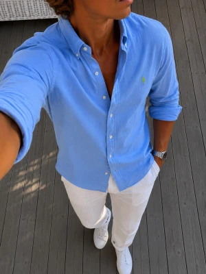 Ralph Lauren skjorta - Snygg blå skjorta med klassisk knappkrage och vita knappar. Skjortan har lång ärm och en liten grön broderad logga på bröstet. Perfekt för en stilren och avslappnad look.