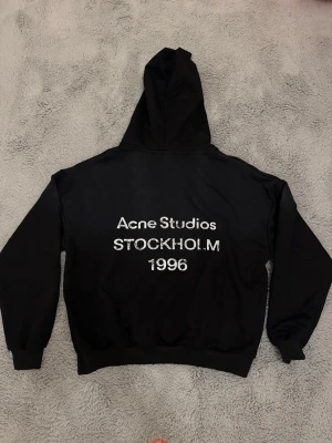 Svart hoodie Acne Studios Stockholm - Säljer en svart hoodie från Acne Studios med tryck på ryggen där det står 'Acne Studios STOCKHOLM 1996'. Klassisk passform med huva och ribbade muddar. Perfekt för dig som gillar stilrena och trendiga plagg med streetwear-vibe.