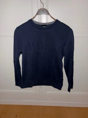 Mörkblå sweatshirt BOSS - Mörkblå långärmad sweatshirt från BOSS med rund halsringning och diskret logotyp framtill. Tröjan har en klassisk passform och är tillverkad i mjukt material som känns skönt mot huden. Perfekt för en avslappnad och stilren look.