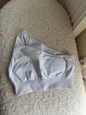 Vit oneshoulder sport-bh - Snygg vit oneshoulder sport-bh med bred ribbad kant nedtill. Bh:n har en sportig look och är tillverkad i mjukt material som känns bekvämt mot huden. Perfekt för dig som gillar unik design och enkel stil.