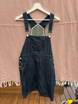 Mörkblå denim jumpsuit med hängslen - Snygg mörkblå denim jumpsuit med klassiska justerbara hängslen och kors i ryggen. Framtill finns en bröstficka och sidofickor samt tre silverfärgade knappar på sidan. Materialet är slitstarkt jeans-tyg i bomull. Perfekt för en avslappnad och trendig look.